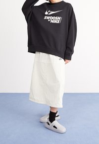 Felpa nera oversize con logo "SWOOSH by NIKE", gonna bianca midi e scarpe grigie senza lacci con ritagli e suola testurizzata.