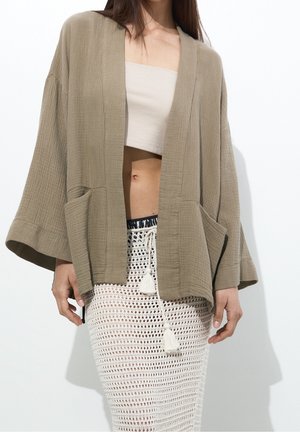 Woman wearing loose beige kimono-style cardigan, cream tube top, and white crochet skirt with drawstring tassels, shown from neck to mid-thigh.