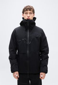 Νέος άνδρας που φοράει μαύρο αδιάβροχο μπουφάν με ψηλό γιακά, πολλαπλές τσέπες με φερμουάρ και ρυθμιζόμενα λουριά στους καρπούς, στέκεται μπροστά σε απλό φόντο.