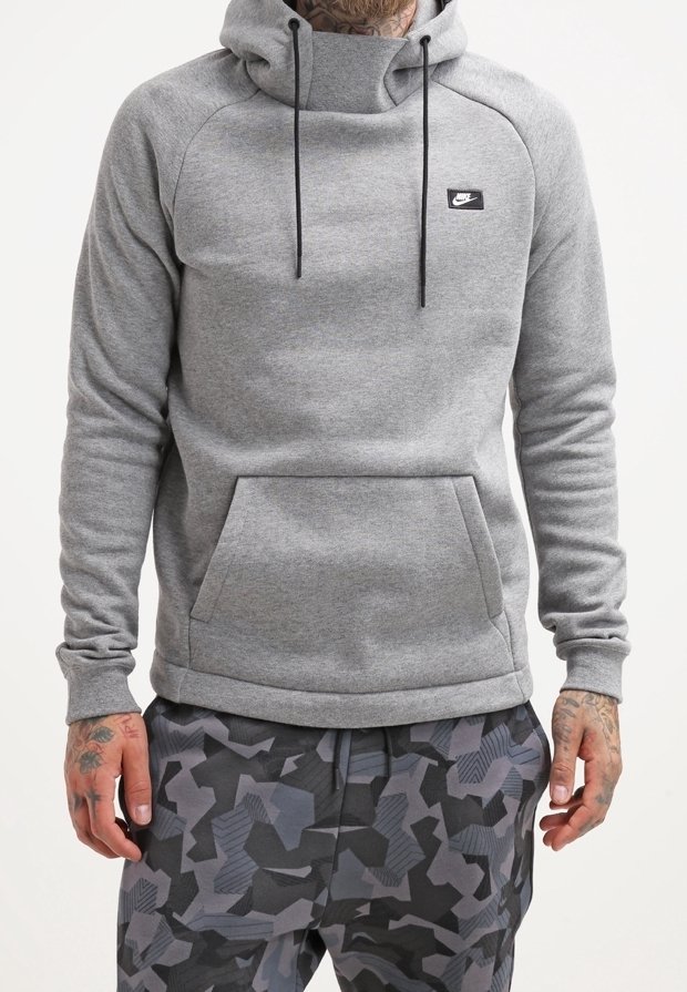 Man in een grijze Nike hoodie met een voorzak en zwart-grijze geometrische camouflagebroek, staand met ontspannen handen langs de zijkanten.