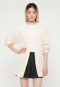 Jeune femme aux cheveux ombrés lisses portant un cardigan en tricot crème sur un haut blanc et une jupe plissée noire, posant avec une main sur la hanche.