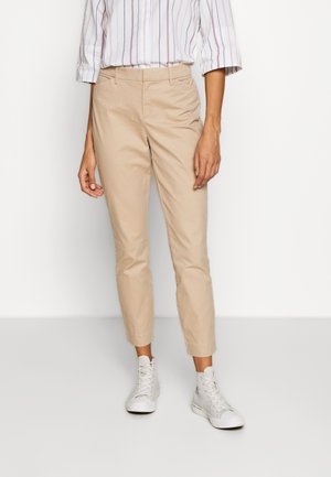 Person trägt beige verkürzte Hose, weiße High-Top-Sneaker und ein weißes Hemd mit dünnen vertikalen Streifen.