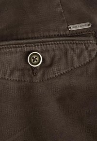 Hnědá látka s texturovaným povrchem, která obsahuje prošívanou kapsu se stříbrným knoflíkem a kovovou visačkou s nápisem "JACK & JONES".