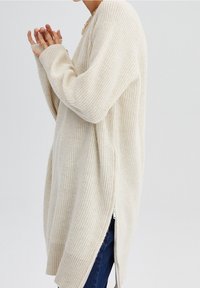 Maglione oversize color crema con una texture a coste, maniche lunghe e cerniere laterali. Abbinato a jeans blu e a una collana semplice.