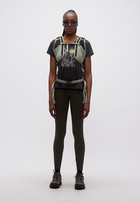 Γυναίκα που φοράει μαύρο μπλουζάκι με γράφικά, πράσινο χακί τσαντάκι στήθους και κολάν, μποτάκια για πεζοπορία και σκούρα γυαλιά ηλίου, στέκεται μπροστά σε λευκό φόντο.