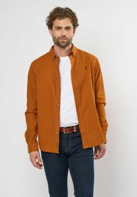 Knoopshirt in roestkleur, gemaakt van een gestructureerde stof met lange mouwen, een kraag en zichtbare knopen. Draag het over een wit t-shirt.