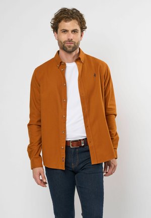 Button-up shirt in rust color, made from a textured fabric with long sleeves, a collar, and visible buttons. Worn over a white t-shirt.
