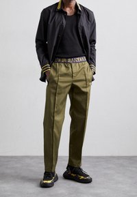 Man wearing olive green trousers, black shirt, black open jacket with yellow trim, and black sneakers with yellow accents on a gray floor.