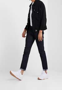 Veste en denim noir avec fermetures à boutons, t-shirt blanc avec motif graphique, jean bleu foncé et baskets blanches avec semelles en caoutchouc. Tenue décontractée.