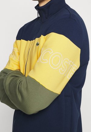 Mann mit einer navy-, gelb- und olivfarbenen Colorblock-Zip-Jacke mit "Lacoste"-Text auf dem gelben Abschnitt und verschränkten Armen.