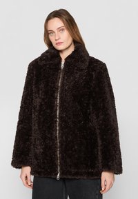 Veste en fausse fourrure marron avec col, fermeture éclair à l'avant et manches longues. Texture douce et design oversize. Modèle se tient contre un fond gris.