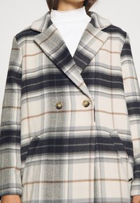 Dubbelt knäppt rutigt rock i kräm, svart och grå nyanser med bruna accenter. Har en notched krage och sidofickor. Mjuk ulltextur.