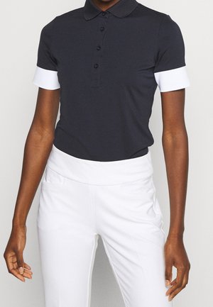 Persoon draagt een zwart poloshirt met korte mouwen en witte manchetten en een witte broek met hoge taille tegen een effen achtergrond.