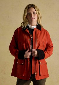 Czerwona wodoodporna kurtka z zamkiem i zatrzaskiem z przodu, brązowe kordowe mankiety, dwie przednie kieszenie i czarny kołnierz. Noszona na wzorzystym swetrze.