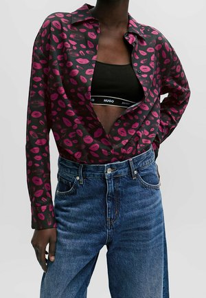 Camicia nera con stampe di labbra rosa, indossata aperta sopra un bralette nero con marchio Hugo, abbinata a jeans di denim blu a vita alta.