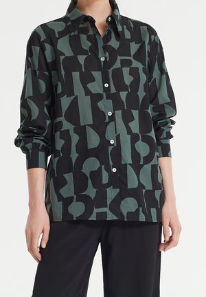 Chemise à manches longues boutonnée avec un motif géométrique abstrait noir et vert foncé, portée avec un pantalon noir.