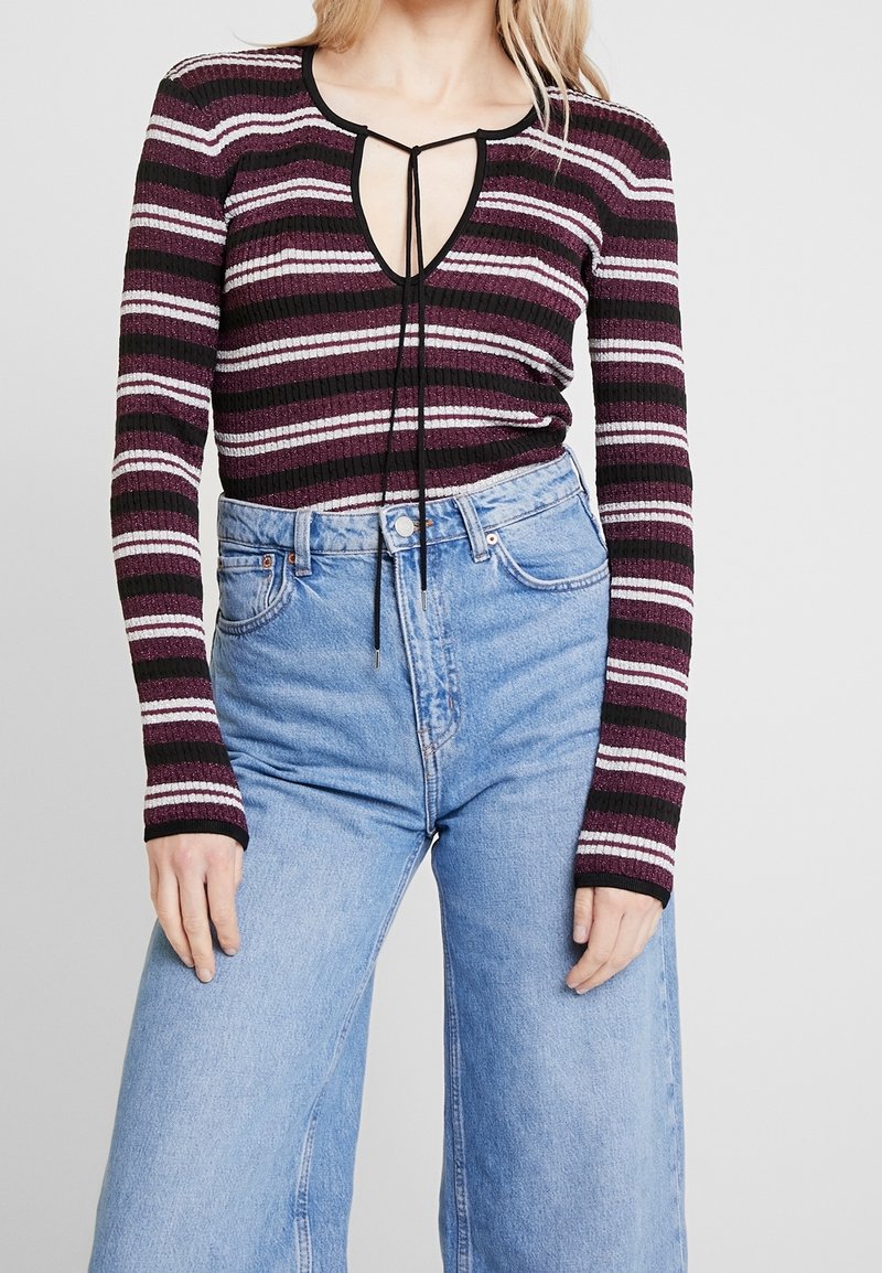 Haut à manches longues rayé avec un profond décolleté en V et un cordon de serrage, présentant des couleurs bordeaux, noir et blanc. Associé à un jean bleu taille haute.