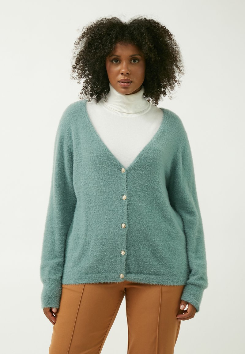 Teal fuzzy cardigan med V-udskæring, fem knapper og lange ærmer, lagdelt over en hvid rullekrave. Blød tekstur og afslappet pasform.