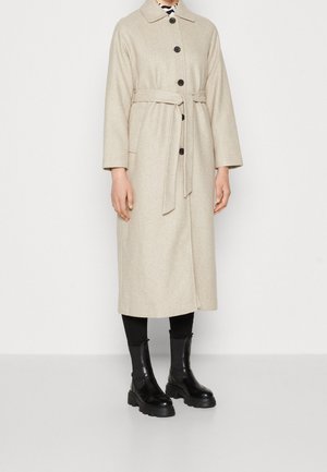 Manteau long en laine beige avec ceinture et boutons noirs, porté sur des leggings noirs et des bottines noires épaisses.