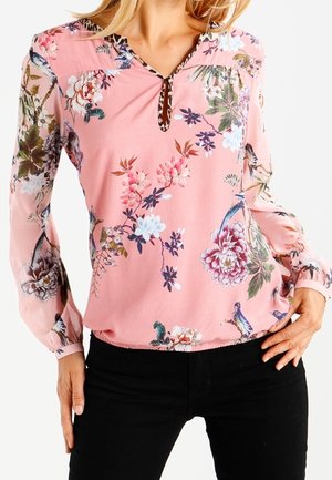 Donna che indossa una camicetta rosa a maniche lunghe con motivi floreali e di uccelli, abbinata a pantaloni neri, in posa con le mani sui fianchi.