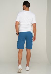 Herren weißes Kurzarmhemd kombiniert mit blauen Cargo-Shorts mit seitlichen Taschen, glatter Textur und lässiger Passform. Weiße Sneaker getragen.