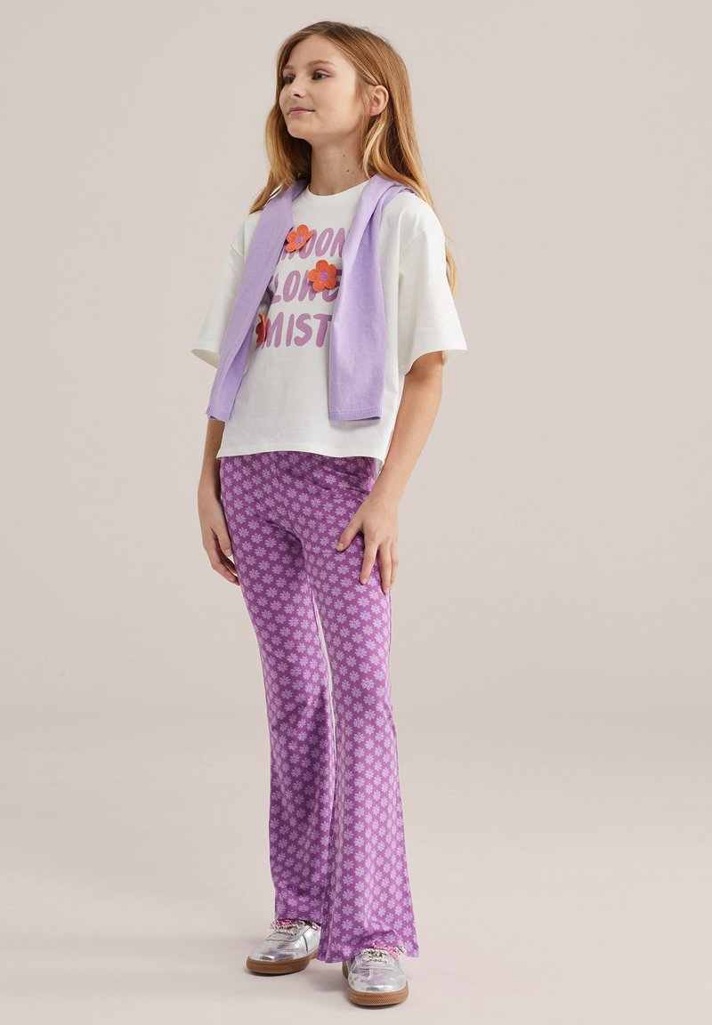 Mergina su ilgais šviesiais plaukais, dėvinti baltą su grafiniu atspaudu marškinėlį, alyvinį megztinį per pečius, purpurinius žieduotus platėjančius kelnes ir sidabrinius sportinius batelius.