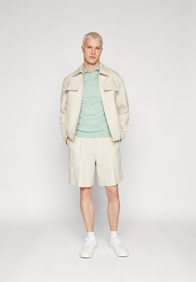 Veste bomber beige sur un polo vert menthe, associée à un short beige. Des baskets blanches apportent du contraste. Textures lisses partout.