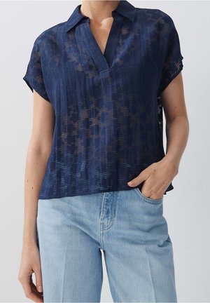 Vrouw draagt een marineblauwe, doorschijnende, gestructureerde blouse met korte mouwen en een gespleten kraag, gecombineerd met lichtblauwe, hooggetaillede jeans, hand in de zak.