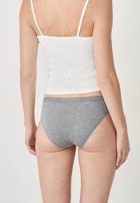 Mujer vistiendo una blusa sin mangas blanca y ropa interior de algodón gris, vista desde atrás contra un fondo blanco liso.