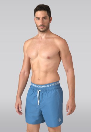 Muž v modrých plaveckých šortkách s pásikom s logom značky a bielou šnúrkou, stojaci na jednoduchom sivom pozadí.