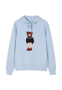 Sudadera de color azul claro con un gráfico de un oso en la parte frontal, detallada con un suéter negro, pantalones beige y toques marrones en los zapatos.