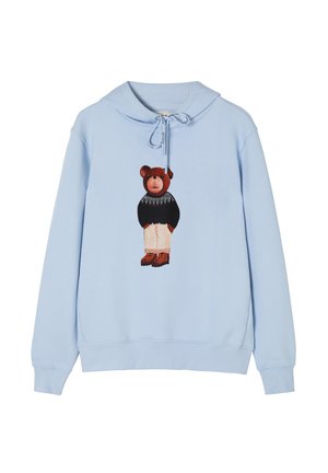 Helles blaues Hoodie mit einem Bärenmotiv auf der Vorderseite, kombiniert mit einem schwarzen Pullover, beigen Hosen und braunen Akzenten an den Schuhen.