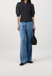 Zwarte gebreide blouse met puffmouwen, knoopsluiting en verticale ribbels; gecombineerd met wijdlopende lichtblauwe spijkerbroek en een zwarte geweven handtas.