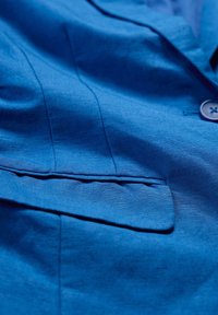 Blå linnakavaj med en ficka, slät textur, subtil lyster och detaljstickning längs sömmarna. Knäppning synlig.