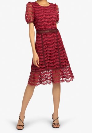 Femme portant une robe en dentelle bordeaux longueur genou avec des manches bouffantes et des sandales noires à talons hauts à brides, debout avec une jambe en avant.