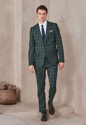 Jeune homme en costume à carreaux vert foncé avec chemise blanche et cravate bleu marine, marchant à l'intérieur sur des marches beige près d'un panier tressé avec une couverture.