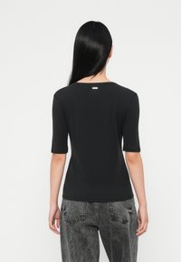 Mujer con cabello largo y negro viste una camiseta negra de manga corta y jeans gris desgastados, de pie de espaldas contra un fondo blanco liso.