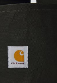 Musta kangastuote, jossa on teksturoidu pinta, varustettuna valkoisella tarralla, jossa on keltainen logo ja "carhartt"-teksti alareunassa.