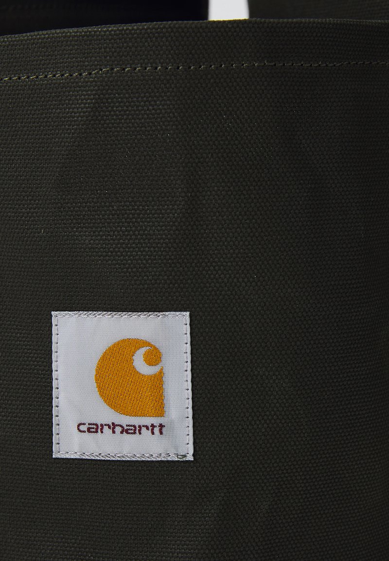 Schwarzes Stoffprodukt mit strukturierter Oberfläche, versehen mit einem weißen Etikett mit gelbem Logo und dem Text "carhartt" am unteren Rand.