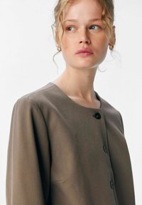 Jeune femme aux cheveux blonds bouclés, portant un manteau taupe boutonné, regardant légèrement sur sa gauche sur un fond blanc.