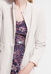 Ljusbeige jacka med mandarinliknande krage, dragkedjestängning och fickdetaljer, bärs över en blommönstrad mörk klänning i rosa och marinblått.