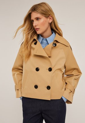 Gabardina beige de doble botonadura con grandes botones negros, solapas anchas y bolsillos frontales, superpuesta a una camisa azul clara.