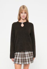 Dunkelbrauner Pullover mit rundem Ausschnitt und Schlüssel-Ausschnitt-Detail, aus weichem Material. Kombiniert mit einem karomuster Minirock in Beige- und Brauntönen.