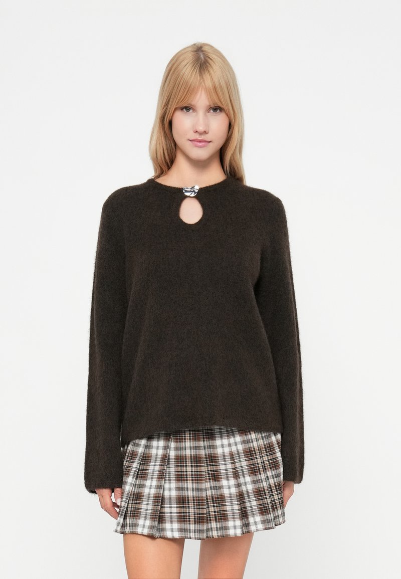Dunkelbrauner Pullover mit rundem Ausschnitt und Schlüssel-Ausschnitt-Detail, aus weichem Material. Kombiniert mit einem karomuster Minirock in Beige- und Brauntönen.