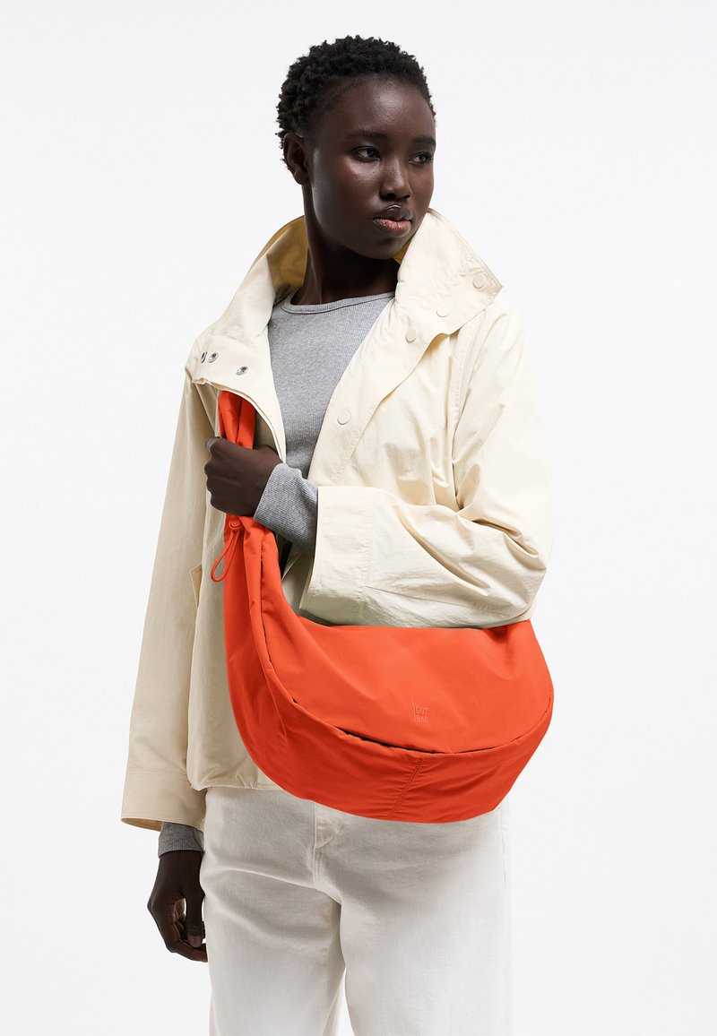 Donna con giacca color crema e camicia grigia tiene una grande borsa a tracolla arancione brillante, guardando leggermente verso destra su sfondo bianco.