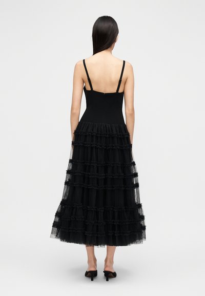 Μαύρο φόρεμα με λεπτές τιράντες, δομημένο μπούστο και πολύφυλλη φούστα από τούλι με βολάν, που φτάνει μέχρι τη μέση της γάμπας. Κλειστή πλάτη.