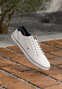 Vita sneakers med marinblå häl och röd, vit och blå kant, svävande över träplattor med små stenar i utomhusmiljö.