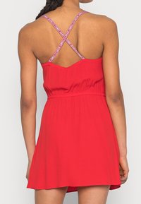 Červené šaty bez rukávů s těsným korzetem, širokou sukní a křížovými látkovými popruhy se zářezem loga. Hladká textura, lehký materiál.