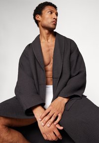 Black waffle-knit robe with wide sleeves, textured fabric, and open front, worn over white underwear. Model seated, arms crossed.
