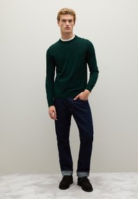 Maglione verde a girocollo realizzato in tessuto a maglia, abbinato a jeans in denim scuri e scarpe casual marroni. Il modello si trova in una posizione contro uno sfondo neutro.